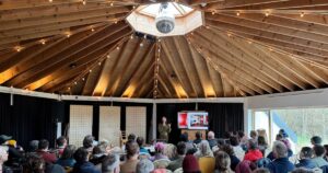 Image of people at an event in an octagon shaped room with someone presenting beside a tv at the front of the room.