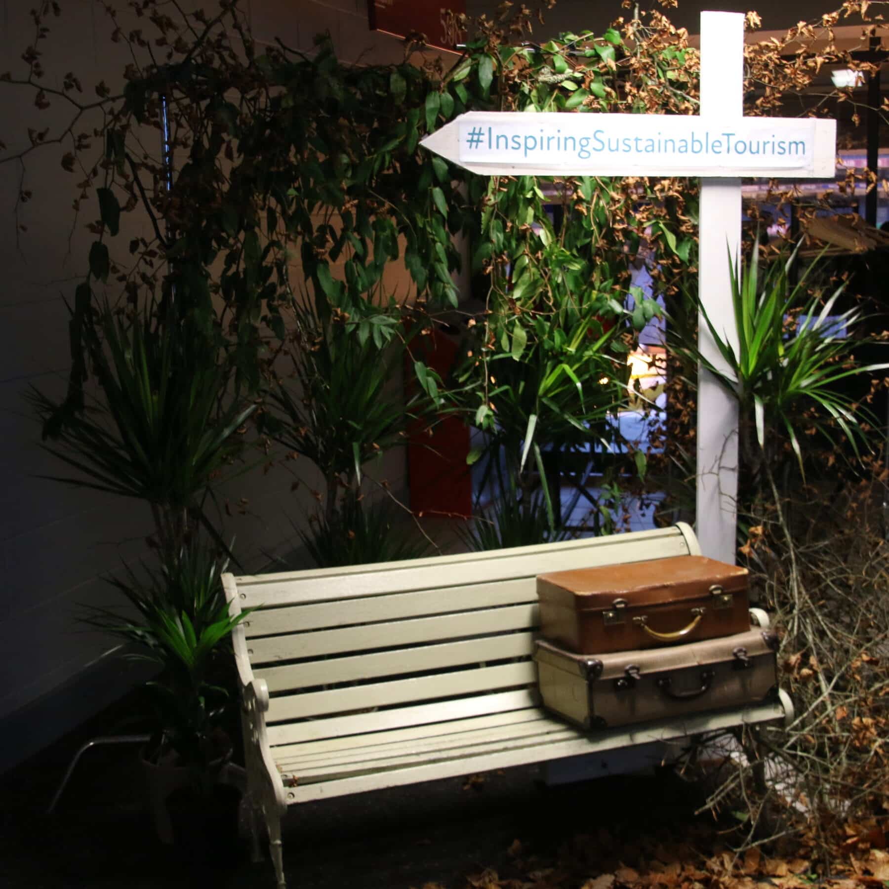 Inspiring Sustainable Tourism. Image of suitcases on white bench with plants behind and white wooden arrow sign saying #InspiringSustainableTourism