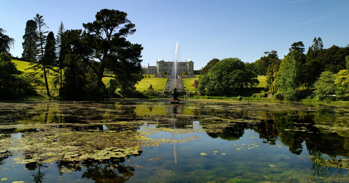 Image of Powerscourt Estate & Gardens. Pond with green vegetation in the foreground. Estate House in the background with green spaces, trees and a water fountain.