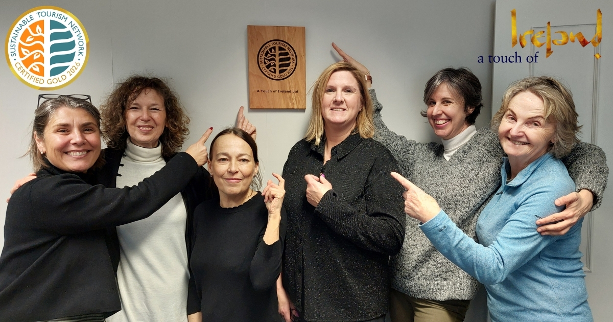 Group of people posing for a photo and smiling while pointing to a sustainable tourism certification wooden plaque. Sustainable Tourism Network Gold Certification logo. A touch of Ireland logo.