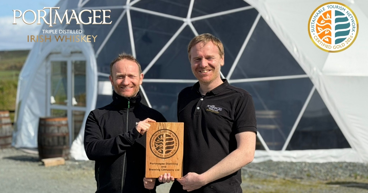 Two people posing for a photo and smiling with a sustainable tourism certification wooden plaque standing outside with black tops on in front of white geo dome sturctures. Sustainable Tourism Network Gold Certification logo. Portmagee Whiskey Triple Distilled Distillery logo